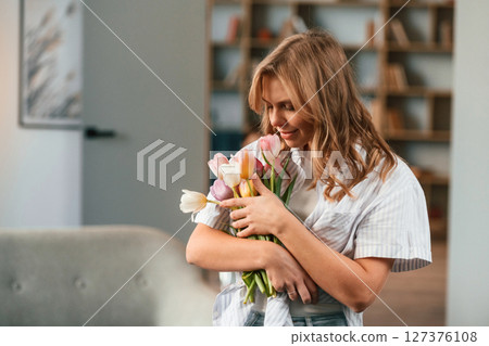 Holding the gift. Young woman is standing in domestic room with flowers 127376108
