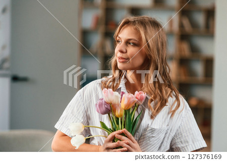 Holding the gift. Young woman is standing in domestic room with flowers 127376169
