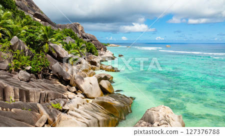 Granite rocks resting on a tropical coastline with turquoise waters and lush green vegetation. La Digue, Seychelles. 127376788