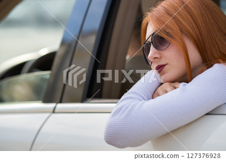 Young woman with red hair and sun glasses travelling by car. Passenger looking out of the rear window of a taxi in a city. Young woman with red hair and sun glasses travelling by car. Passenger looking out of the rear window of a taxi in a city. 127376928