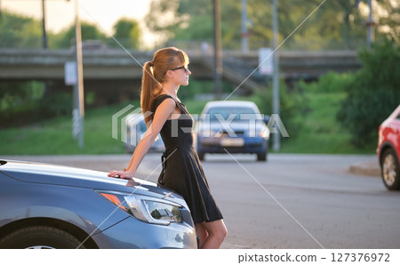 Young woman driver enjoying warm summer day standing beside her car on city street. Travelling and vacation concept Young woman driver enjoying warm summer day standing beside her car on city street. Travelling and vacation concept 127376972