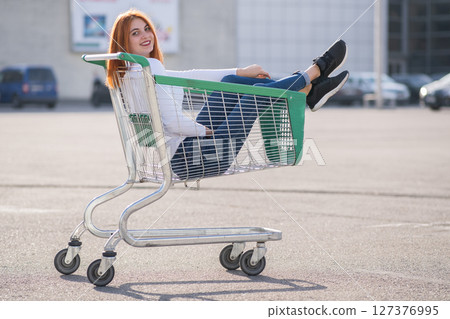 Young teenage happy girl sitting in supermarket shopping cart outdoors. 127376995