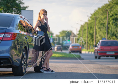 Young female driver standing near her car talking on mobile phone on a city street in summer 127377041