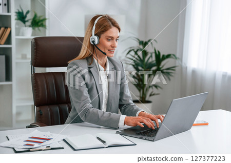 In headphones, by laptop, call center. Woman in business formal clothes is working in office 127377223