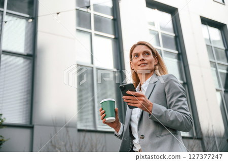 With cup of drink and phone. Woman in formal clothes is outdoors near the business building 127377247