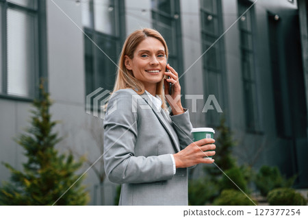 Walking, holding cup of drink and talking by phone. Woman in formal clothes is outdoors near the business building Walking, holding cup of drink and talking by phone. Woman in formal clothes is outdoors near the business building 127377254