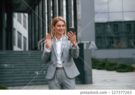 Online call by using smartphone. Woman in formal clothes is outdoors near the business building Online call by using smartphone. Woman in formal clothes is outdoors near the business building 127377263