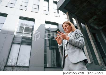 View from below. Walking with smartphone. Woman in formal clothes is outdoors near the business building View from below. Walking with smartphone. Woman in formal clothes is outdoors near the business building 127377271