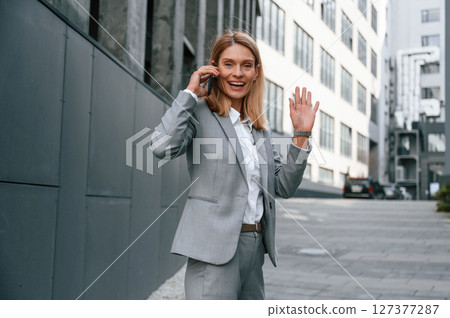 Conversation by the phone call. Woman in formal clothes is outdoors near the business building Conversation by the phone call. Woman in formal clothes is outdoors near the business building 127377287
