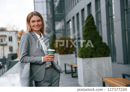 Smiling, holding cup of drink. Woman in formal clothes is outdoors near the business building Smiling, holding cup of drink. Woman in formal clothes is outdoors near the business building 127377289