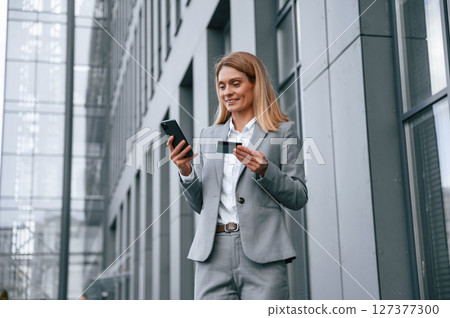 Holding credit card and smartphone. Woman in formal clothes is outdoors near the business building 127377300