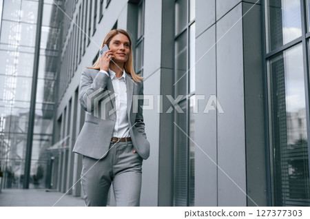 Conversation by the phone. Woman in formal clothes is outdoors near the business building Conversation by the phone. Woman in formal clothes is outdoors near the business building 127377303