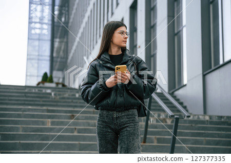 Standing on the stairs. Young woman is outdoors near building is holding smartphone Standing on the stairs. Young woman is outdoors near building is holding smartphone 127377335