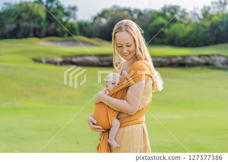 Mother holding her little baby in a yellow sling in the park. Warm and loving family moment. Babywearing, parenting, and mother-child bonding concept 127377386