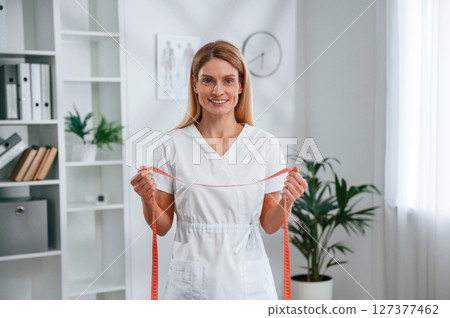 With measuring tape in hands, conception of diet. Young female doctor in white coat is indoors With measuring tape in hands, conception of diet. Young female doctor in white coat is indoors 127377462