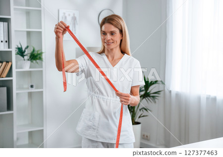 With measuring tape in hands, conception of diet. Young female doctor in white coat is indoors With measuring tape in hands, conception of diet. Young female doctor in white coat is indoors 127377463
