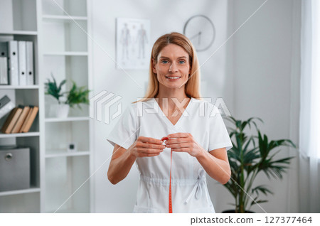 With measuring tape in hands, conception of diet. Young female doctor in white coat is indoors 127377464