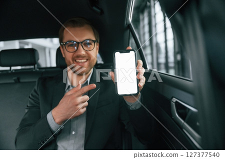 Holding smartphone. Man in formal business clothes is sitting in the modern automobile 127377540