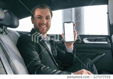 Holding smartphone. Man in formal business clothes is sitting in the modern automobile 127377559
