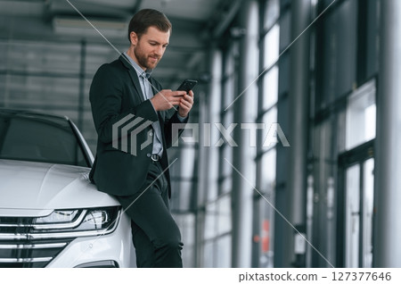 Standing and holding smartphone. Man in black suit is indoors in the car dealership near automobile 127377646
