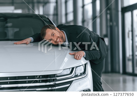 Embracing the vehicle. Man in black suit is indoors in the car dealership near automobile Embracing the vehicle. Man in black suit is indoors in the car dealership near automobile 127377650