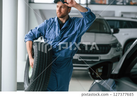 Walking forward and holding the tire. Man in blue uniform is working in the car service Walking forward and holding the tire. Man in blue uniform is working in the car service 127377685