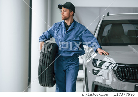 With tire in the hands. Man in blue uniform is working in the car service 127377686