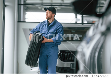 With tire in the hands. Man in blue uniform is working in the car service With tire in the hands. Man in blue uniform is working in the car service 127377688