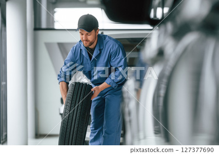 With tire in the hands. Man in blue uniform is working in the car service With tire in the hands. Man in blue uniform is working in the car service 127377690