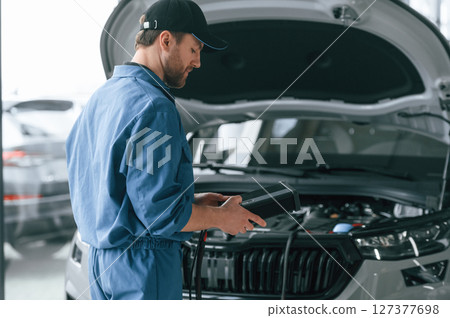 Holding electricity measuring tool. Man in blue uniform is working in the car service Holding electricity measuring tool. Man in blue uniform is working in the car service 127377698