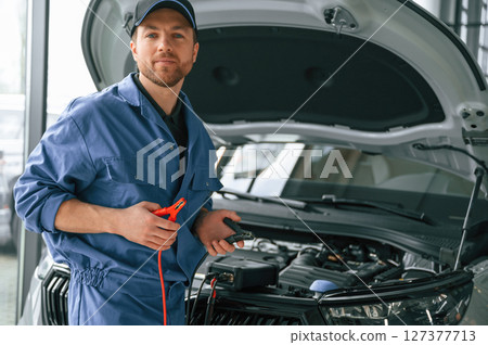 Holding the electrical terminals. Man in blue uniform is working in the car service 127377713