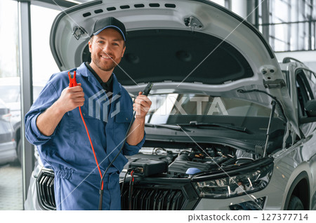 Holding the electrical terminals. Man in blue uniform is working in the car service 127377714