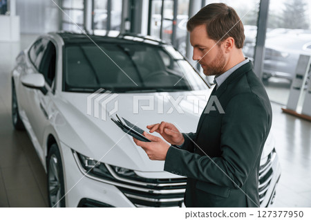 Standing, side view, holding tablet. Man in black suit is indoors in the car dealership near automobile Standing, side view, holding tablet. Man in black suit is indoors in the car dealership near automobile 127377950
