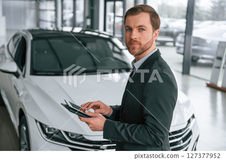 Standing, side view, holding tablet. Man in black suit is indoors in the car dealership near automobile 127377952