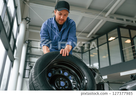 Front view, moving the tire. Man in blue uniform is working in the car service Front view, moving the tire. Man in blue uniform is working in the car service 127377985