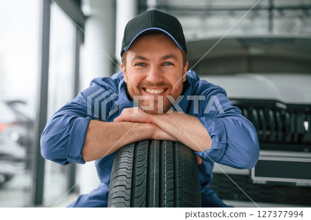 Leaning on the tire and smiling. Man in blue uniform is working in the car service 127377994