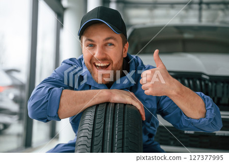 Leaning on the tire and smiling. Man in blue uniform is working in the car service 127377995