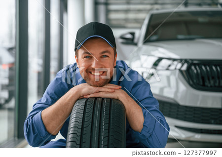 Leaning on the tire and smiling. Man in blue uniform is working in the car service Leaning on the tire and smiling. Man in blue uniform is working in the car service 127377997