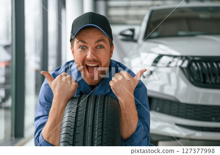 Leaning on the tire and smiling. Man in blue uniform is working in the car service 127377998