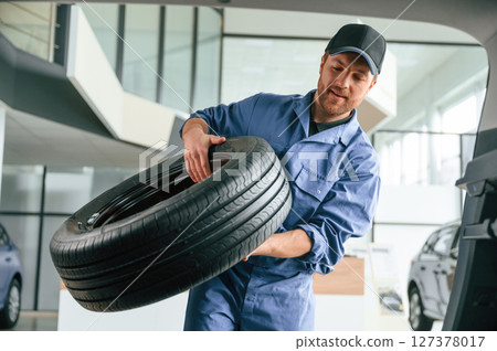With big new tire for a change. Man in blue uniform is working in the car service 127378017