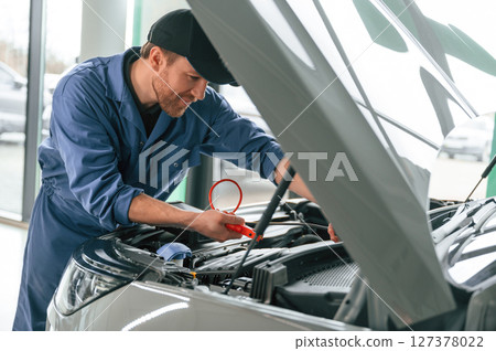 With hood opened. Man in blue uniform is working in the car service With hood opened. Man in blue uniform is working in the car service 127378022