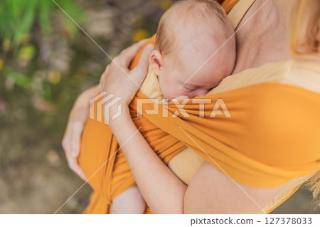 Mother holding her little baby in a yellow sling in the park. Warm and loving family moment. Babywearing, parenting, and mother-child bonding concept Mother holding her little baby in a yellow sling in the park. Warm and loving family moment. Babywearing, parenting, and mother-child bonding concept 127378033