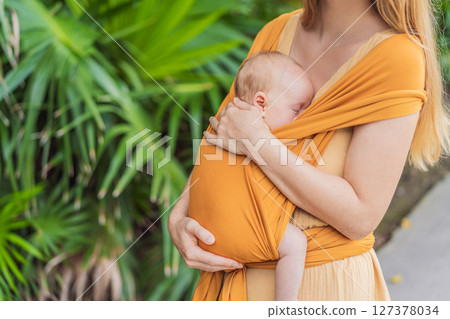 Mother holding her little baby in a yellow sling in the park. Warm and loving family moment. Babywearing, parenting, and mother-child bonding concept 127378034