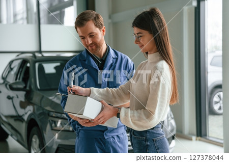 Man is holding box and notepad. Young woman is visiting auto mechanic in garage. Repair service 127378044