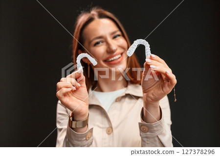 Smiling, and holding two aligners. Young woman is standing against black background 127378296