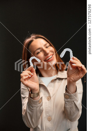 Smiling, and holding two aligners. Young woman is standing against black background Smiling, and holding two aligners. Young woman is standing against black background 127378298