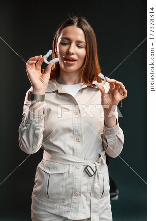 Holding aligner and looking at it. Young woman is standing against black background 127378314