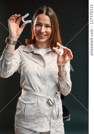 Positive young woman is standing against black background and holding aligner 127378315