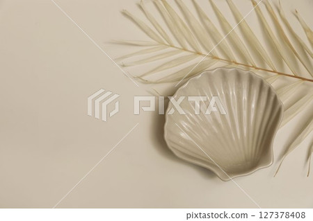 Tropical summer still life. Ceramic seashell plate, dry palm leaf on beige table background. Neutral tones, soft lighting, empty copy space. Sea coastal, minimalistic web banner. Flat lay, top view 127378408