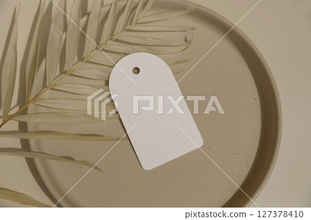 Empty round gift tag, label on speckled ceramic plate in sunlight. Beige table background. Dry palm leaf. Shadows. Elegant summer sale, business template, web banner. Flat lay, top view. Copy space. 127378410